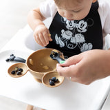 A toddler in a high chair eats from a bowl, assisted by an adult hand with a Bumkins Silicone Dipping Spoons: Mickey Mouse (Classic Black).