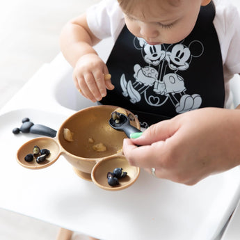 A toddler in a high chair eats from a bowl, assisted by an adult hand with a Bumkins Silicone Dipping Spoons: Mickey Mouse (Classic Black).