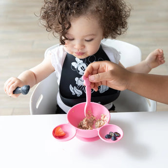 A toddler sits in a high chair, while an adult feeds them with a Bumkins Silicone Dipping Spoons: Minnie Mouse (Black and Pink) from a bowl.