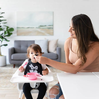 A toddler in a high chair wears a bib while an adult feeds them with a Bumkins Silicone Dipping Spoons: Minnie Mouse (Black and Pink).