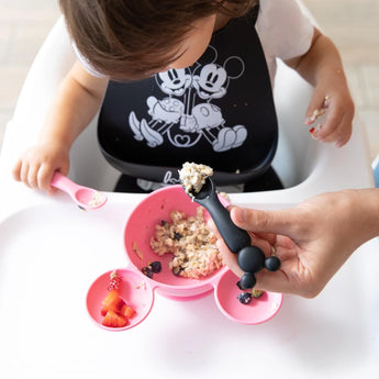 A toddler in a high chair is fed by an adult hand with a Bumkins Silicone Dipping Spoons: Minnie Mouse (Black and Pink) from a pink bowl.