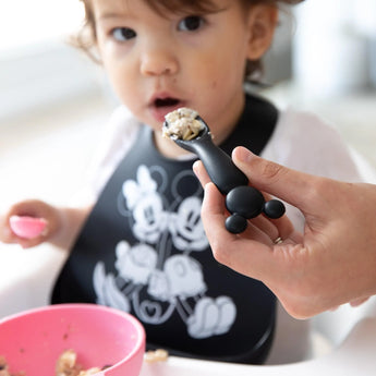 A toddler in a bib is fed by an adult hand with a Bumkins Silicone Dipping Spoons: Minnie Mouse (Black and Pink), while a bowl sits nearby.