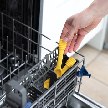 Hands place Bumkins Silicone Dipping Spoons: Winnie the Pooh in the utensil compartment of an open dishwasher.