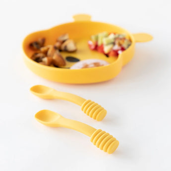 Bumkins Silicone Dipping Spoons: Winnie the Pooh sit on a white background, with a blurred bear-shaped plate of food behind.