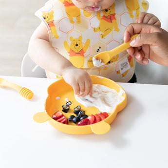 A baby in a bib reaches for fruit and yogurt on a yellow bear plate, aided by an adult with Bumkins Silicone Dipping Spoons: Winnie the Pooh.