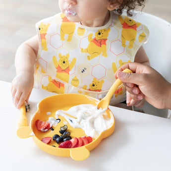 A child enjoys yogurt with fruit on a bear-shaped plate, using Bumkins Silicone Dipping Spoons: Winnie the Pooh with adult assistance.
