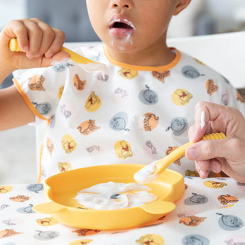 A young child in a colorful bib eats from a plate with Bumkins Silicone Dipping Spoons: Winnie the Pooh, while an adult assists.