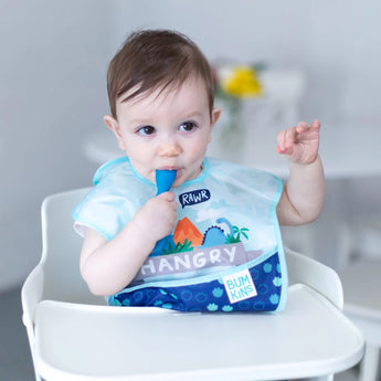 A baby in a high chair, wearing a dinosaur bib, chews a dark blue Bumkins Silicone Dipping Spoons 3 Pack: Gumdrop.