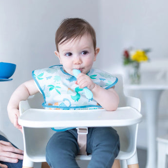 A baby in a high chair wearing a dinosaur bib holds a blue Bumkins Silicone Dipping Spoons 3 Pack: Gumdrop near their mouth.