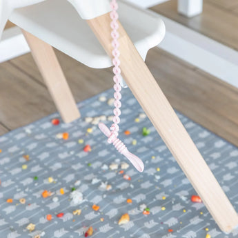 A tether attached to a high chair holds a Bumkins Silicone Dipping Spoons 3 Pack: Lollipop, dangling above a floor mat covered with food.