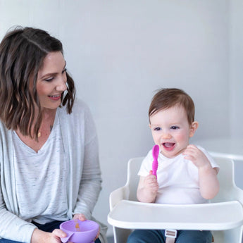 A woman holding a purple bowl smiles at a baby in a high chair holding a Bumkins Silicone Dipping Spoons 3 Pack: Lollipop.