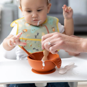 A baby in a bib grabs a Bumkins Silicone Dipping Spoons 3 Pack: Rocky Road in a high chair, with an adult uses another to feed from a bowl.