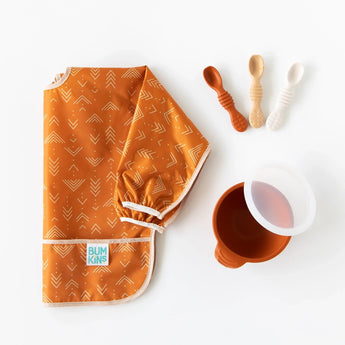 A folded orange bib, a bowl with a transparent lid, and Bumkins Silicone Dipping Spoons 3 Pack: Rocky Road sit on a white background.