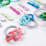 Bumkins Silicone Teething Charms: Pink lays next to other teething toys and silicone spoons on a white marble background.