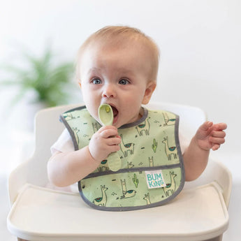 A baby in a green llama bib sits in a high chair, placing a green Bumkins Silicone Dipping Spoons 3 Pack: Taffy in their mouth.