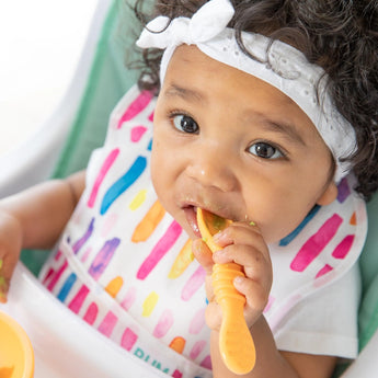 A baby with curls, wearing a white headband and colorful bib, chews a yellow Bumkins Silicone Dipping Spoons 3 Pack: Tutti-Frutti.