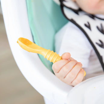 Close-up of a baby in a patterned bib in a high chair, holding a yellow Bumkins Silicone Dipping Spoons 3 Pack: Tutti-Frutti.