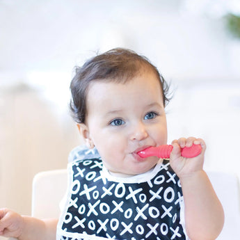 A curly-haired baby in a black and white bib chews a red Bumkins Silicone Dipping Spoons 3 Pack: Tutti-Frutti.