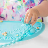 Close-up of a child's hand holding a Bumkins Silicone Chewtensils®: Blue on a blue silicone placemat with cereal pieces.