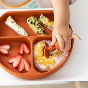 Close-up of a child's hand reaching for yogurt from a divided plate on a high chair tray with Bumkins Silicone Chewtensils®: Clay.