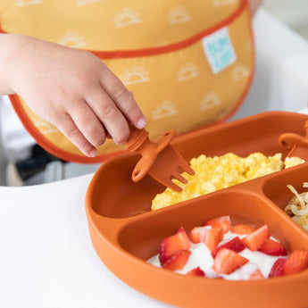 Close-up of a child using Bumkins Silicone Chewtensils®: Clay to pick up eggs from a divided plate, with strawberries and yogurt nearby.