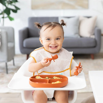 A toddler with pigtails in a high chair wearing a beige bib uses Bumkins Silicone Chewtensils®: Clay to eat from a divided plate.