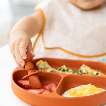 Close-up of a child using Bumkins Silicone Chewtensils®: Clay to pick up strawberries from a divided plate with scrambled eggs and yogurt.