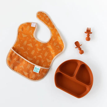 Bumkins Silicone Chewtensils®: Clay sits next to a matching plate and an orange bib with an abstract print on a white background.