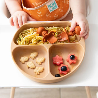 A baby uses Bumkins Silicone Chewtensils®: Clay to eat from a plate with pasta, fruit flowers, and animal crackers on a high chair tray.