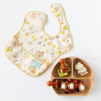 A brown divided plate of food with Bumkins Silicone Chewtensils®: Clay sits next to a colorful camping-themed bib on a white background.