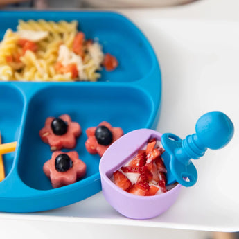 Close-up of a blue plate with pasta and fruit, with a Bumkins Silicone Chewtensils®: Dark Blue in a purple cup with berries and yogurt.