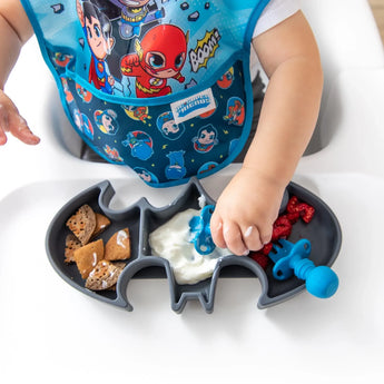 A baby in a high chair wearing a bib reaches for food on a bat-shaped plate with snacks, using Bumkins Silicone Chewtensils®: Dark Blue.