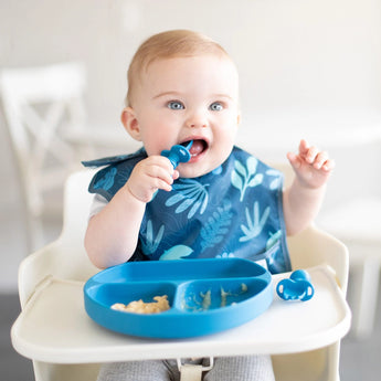 A baby in a high chair wearing a blue bib uses Bumkins Silicone Chewtensils®: Dark Blue to eat from a matching plate.