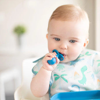 A baby in a high chair, wearing a dinosaur bib, looks intently at a blue plate while chewing a Bumkins Silicone Chewtensils®: Dark Blue.