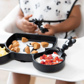 Close-up of a child in a high chair holding Bumkins Silicone Chewtensils®: Mickey Mouse with a matching plate with breakfast foods.