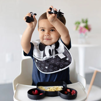 A toddler in a high chair holds Bumkins Silicone Chewtensils®: Mickey Mouse above their head while a black plate of food sits on the tray.