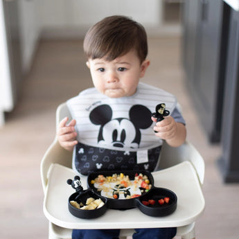 A toddler wearing a black & white bib in a high chair uses Bumkins Silicone Chewtensils®: Mickey Mouse to eat from a Mickey-shaped plate.
