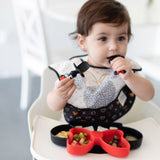 A toddler in a high chair chews Bumkins Silicone Chewtensils®: Minnie Mouse with a Minnie Mouse-shaped plate with food on the tray.