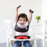 A baby in a high chair happily raises their arms, holding Bumkins Silicone Chewtensils: Minnie Mouse, with a black & red plate nearby.