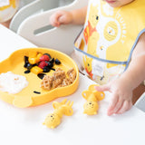 A child in a high chair with a yellow bib reaches for Bumkins Silicone Chewtensils®: Winnie The Pooh next to a bear-shaped plate with food.
