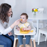 A woman feeds baby in a high chair wearing yellow bib with Bumkins Silicone Chewtensils®: Winnie The Pooh; a table with flowers sits behind.