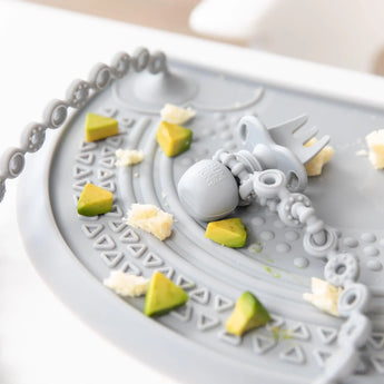Close-up of a Bumkins Silicone Chewtensils®: Gray attached to a gray tether on a gray placemat with avocado pieces.