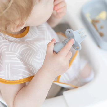 Close-up of a toddler in a high chair wearing a beige bib and holding a Bumkins Silicone Chewtensils®: Gray over a gray plate.
