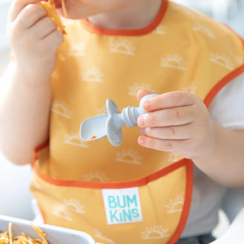 Close-up of a child wearing a yellow bib holding a Bumkins Silicone Chewtensils®: Gray.