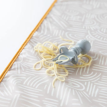 Close-up of a Bumkins Silicone Chewtensils®: Gray with spaghetti on a beige waterproof mat on a white background.