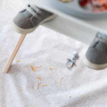 A Bumkins Silicone Chewtensils®: Gray sits on a beige mat with spaghetti under a high chair, with toddler shoes blurred in the foreground