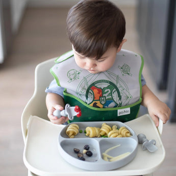 A toddler in a high chair wearing a green bib enjoys fruit and pasta from a gray plate with Bumkins Silicone Chewtensils®: Gray.