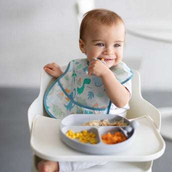 A baby wearing a dinosaur bib in a high chair uses Bumkins Silicone Chewtensils®: Gray to explore baby-led weaning from a gray plate.