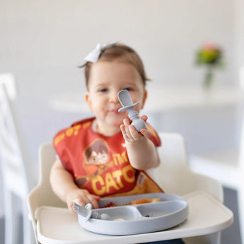 A baby wearing a red bib in a high chair holds a Bumkins Silicone Chewtensils®: Gray toward the camera with a gray plate on the tray.