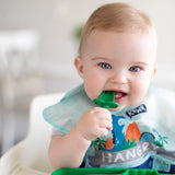 A baby with light hair in a dinosaur bib chews a Bumkins Silicone Chewtensils®: Jade in a high chair.